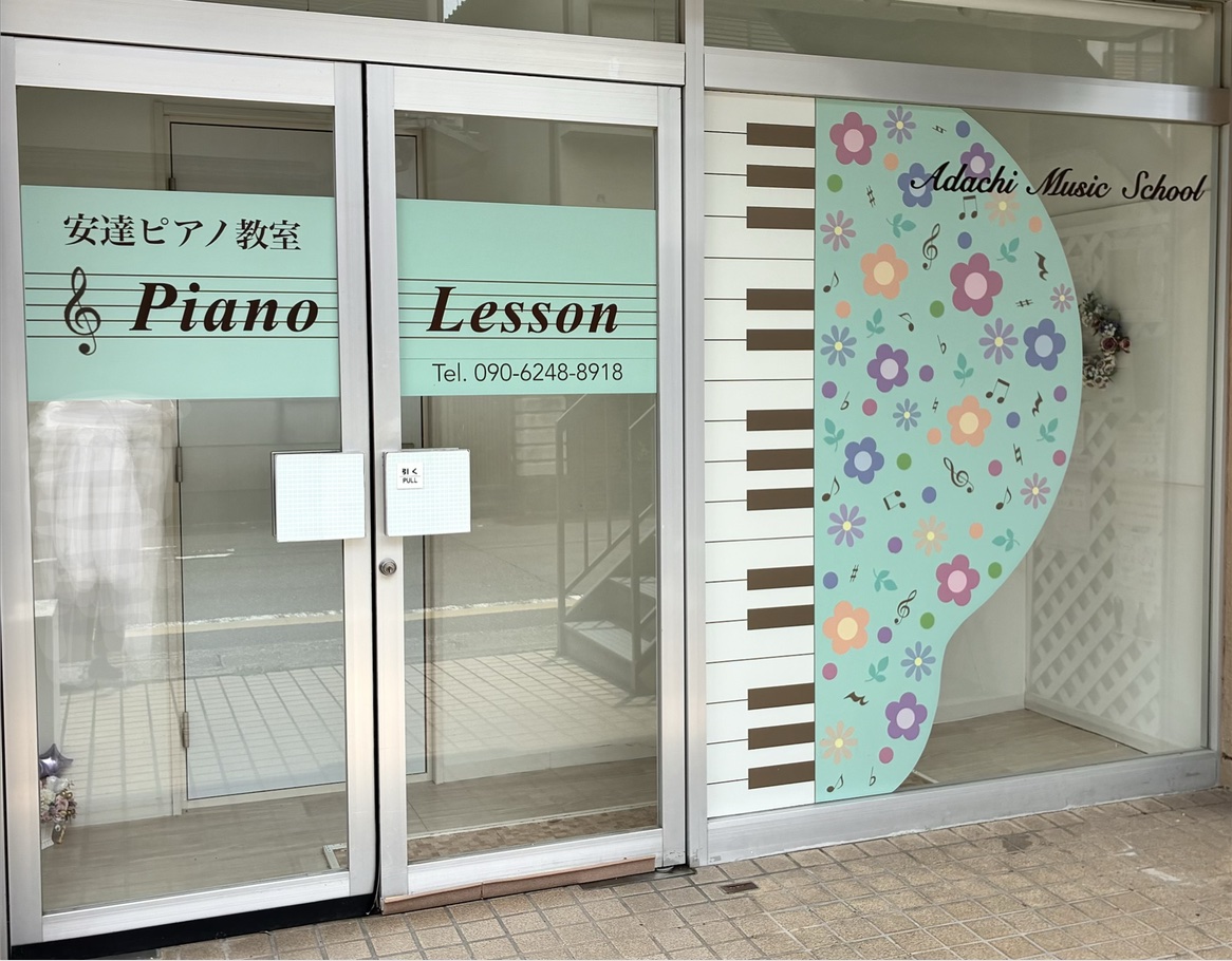 安達ピアノ教室の入り口。ガラス戸に「安達ピアノ教室 Piano Lesson」の看板と、鍵盤と花のかざりがある